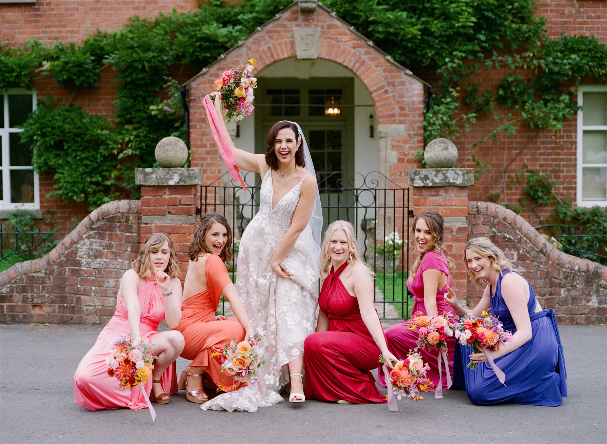 A film photo of bride in a lace dress with bridesmaids in colourful gowns holding bouquets in front of a brick building.