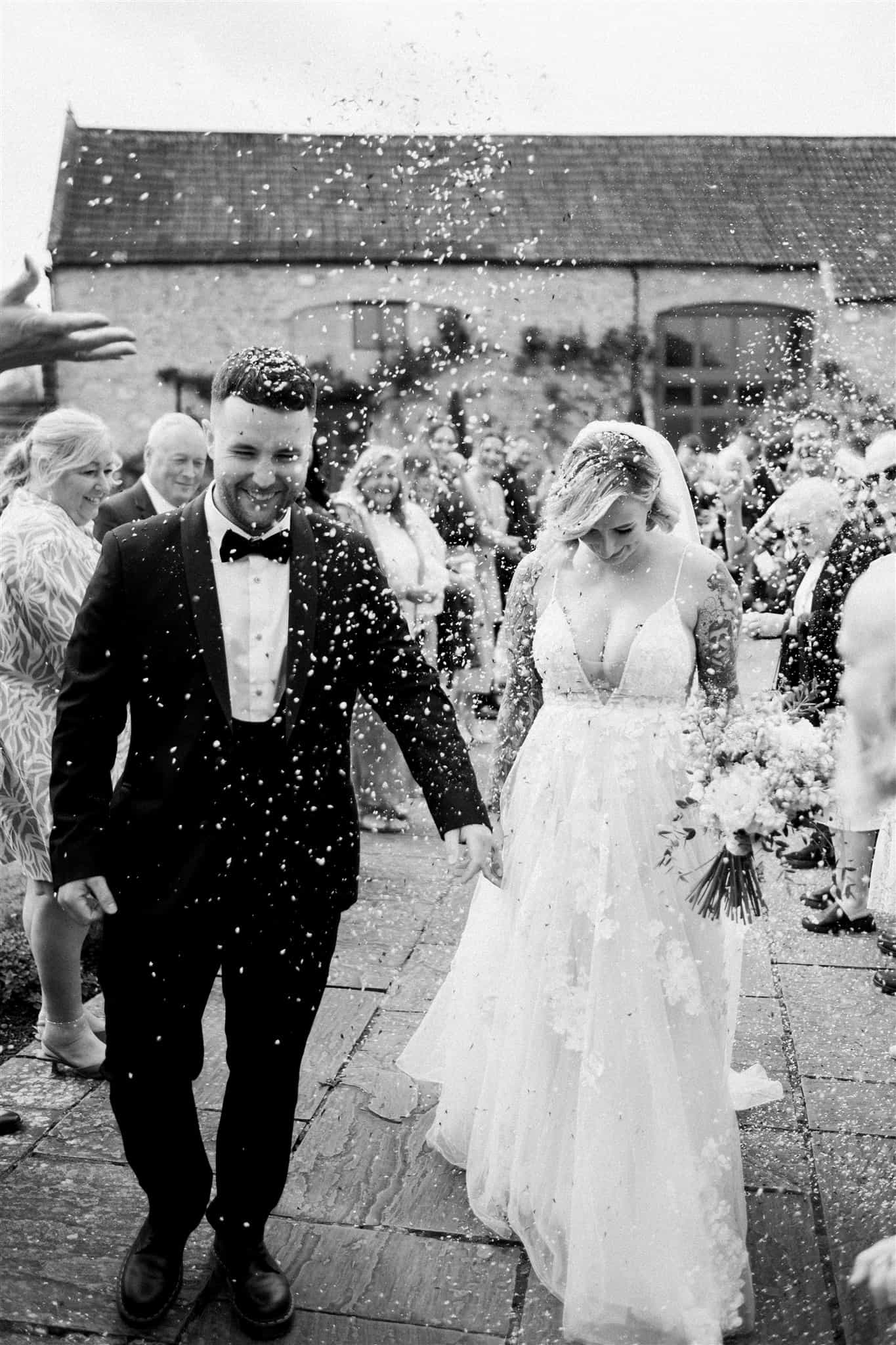 Joyful couple walks down the tunnel, enveloped in confetti at their Priston Mill wedding, photographed by Ivy and Pine.